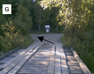 Waypoint G: the view from a small plank bridge, with an arrow pointing left to the road climbing away just past the far end.